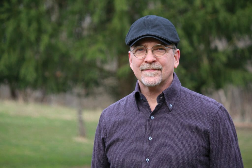 Stan Stewart with a blue cap on and a serious expression. There's a forest in the background. (Don't worry, it's easy to get to know Stan.)