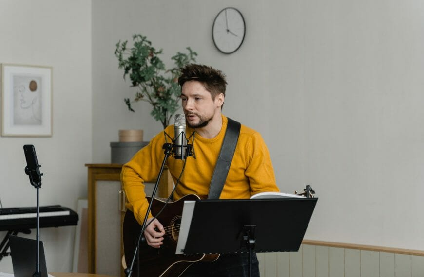 Work at music - Wearing a bright-colored, long-sleeve shirt, a man with a beard and dark hair plays a guitar while singing into a microphone