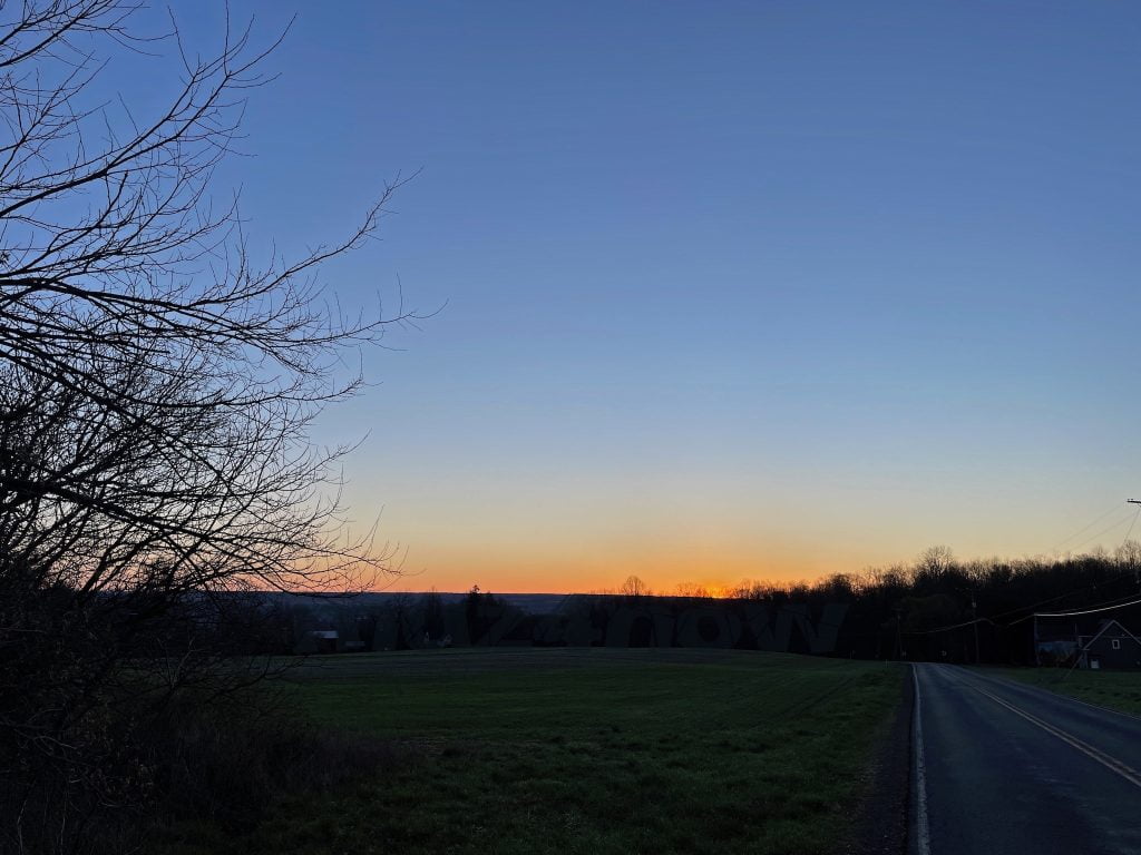 A blue, cloud-free sky with the sun rising on the horizon. A leafless tree is off to the left.