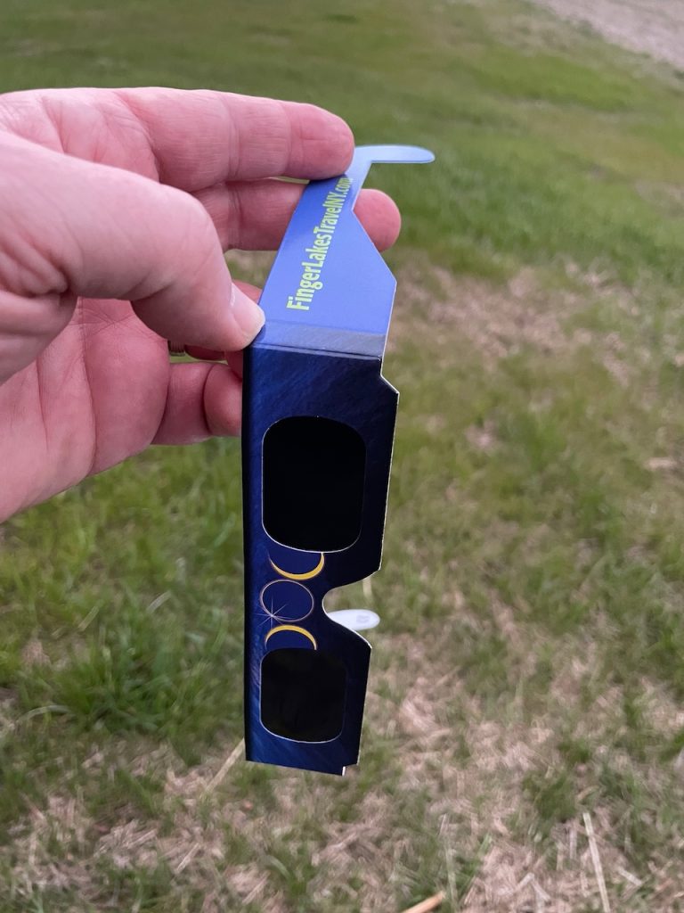 A human hand holding a pair of eclipse glasses lightly. The glasses have "Finger Lakes Travel NY" printed on the side.