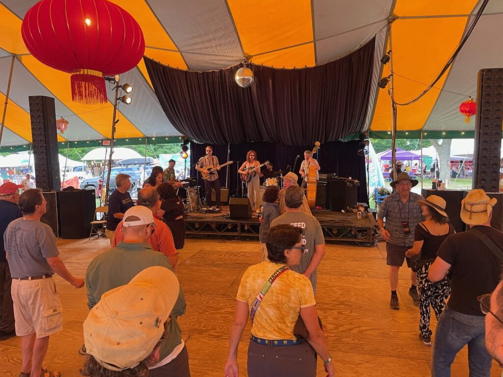 A four-piece band plays while people dance and listen in a yellow and gray tent.