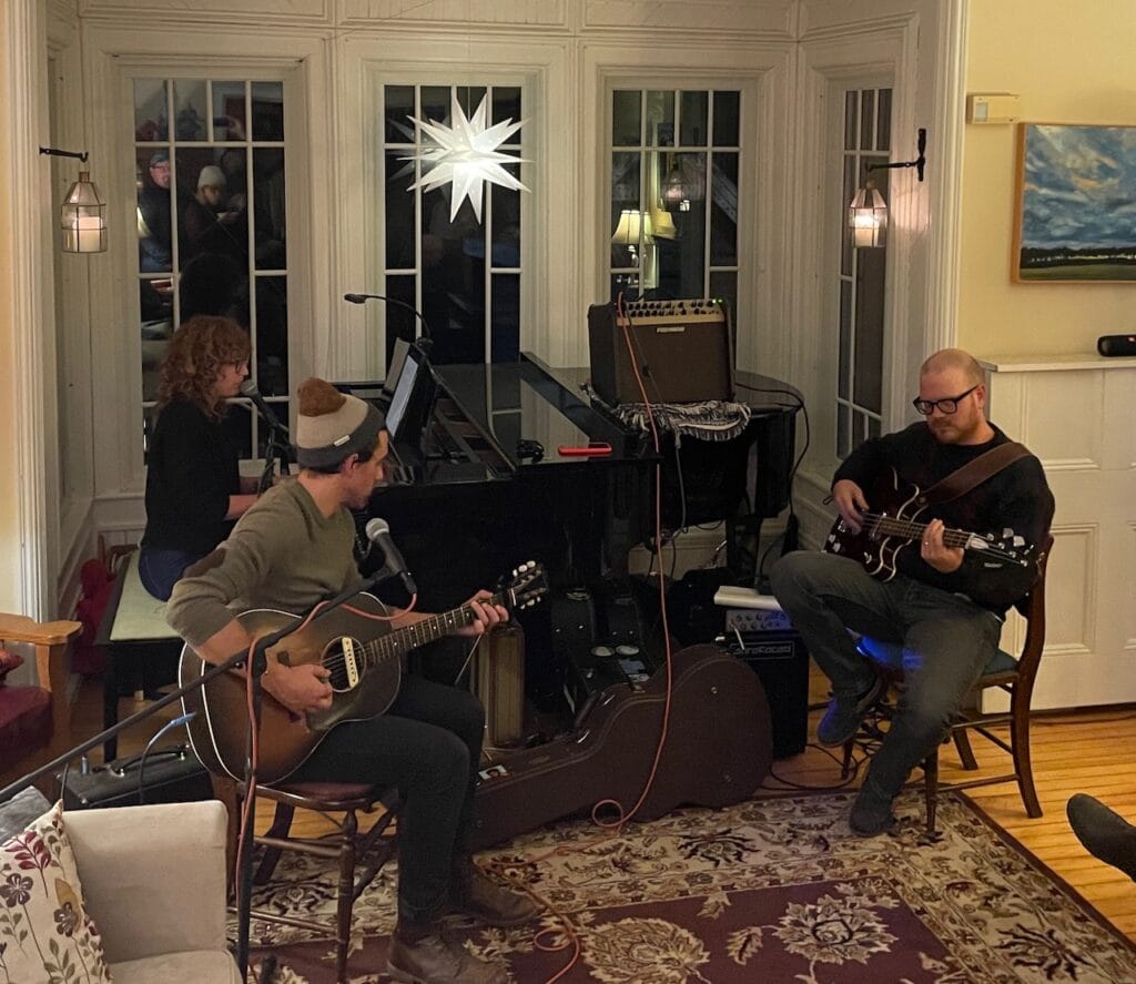 The band Louiston playing a live gig. Louis plays an acoustic guitar. He's sitting on the left side. On the right is Chris playing a semi-hollow bass guitar. Colleen is behind Lewis at an acoustic piano. There's a bright Bethlehem star in the window behind them.