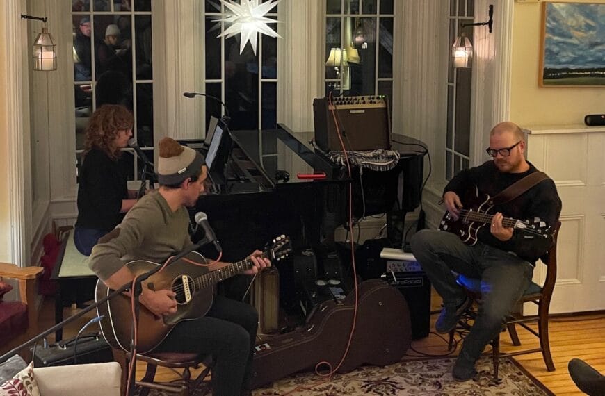 The band Louiston playing a live gig. Louis plays an acoustic guitar. He's sitting on the left side. On the right is Chris playing a semi-hollow bass guitar. Colleen is behind Lewis at an acoustic piano. There's a bright Bethlehem star in the window behind them.