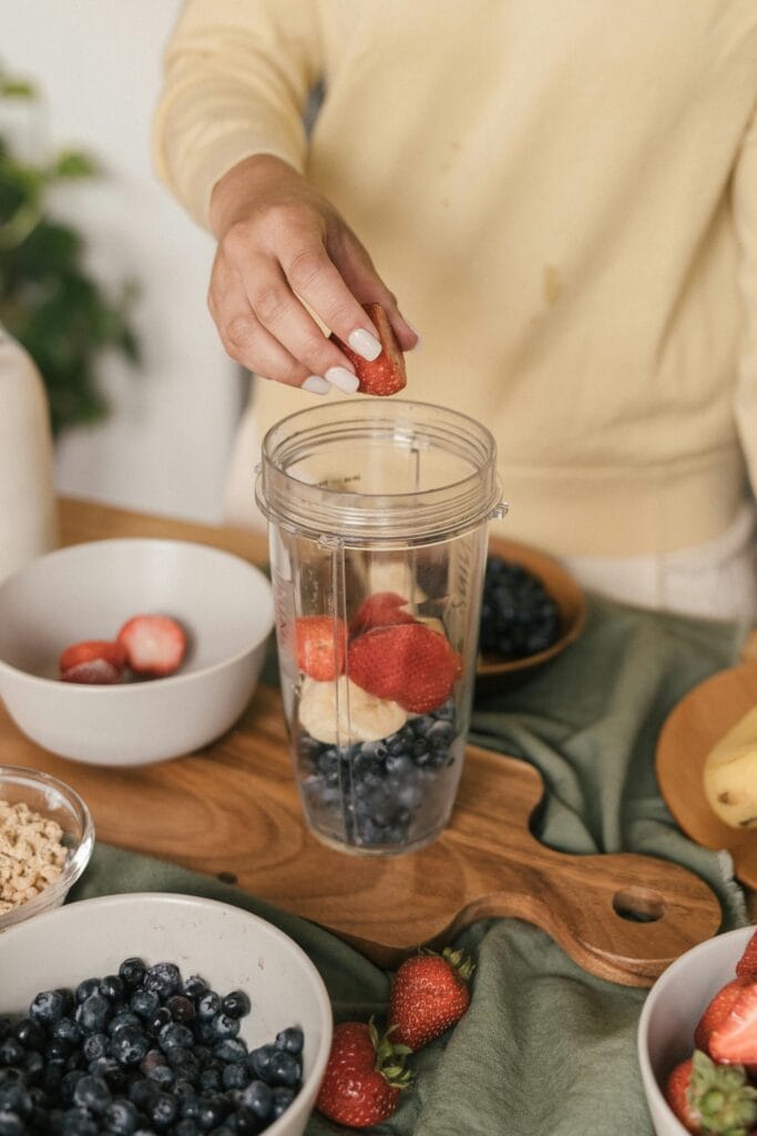 A person with painted fingernails adds fruit to a a clear container with a threaded top for putting in a blender. There are bowls of fruit on the table around the clear container. Explore creating food.
Image by Mike Jones via pexels.