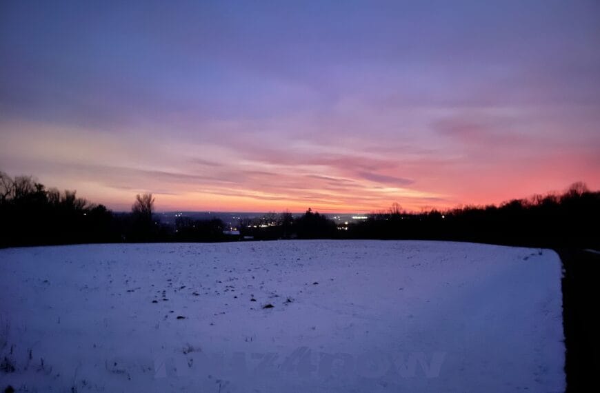 Ready for beauty? Here's a red, orange, and yellow hue on the horizon as the sun rises behind the clouds. The ground is covered with snow.