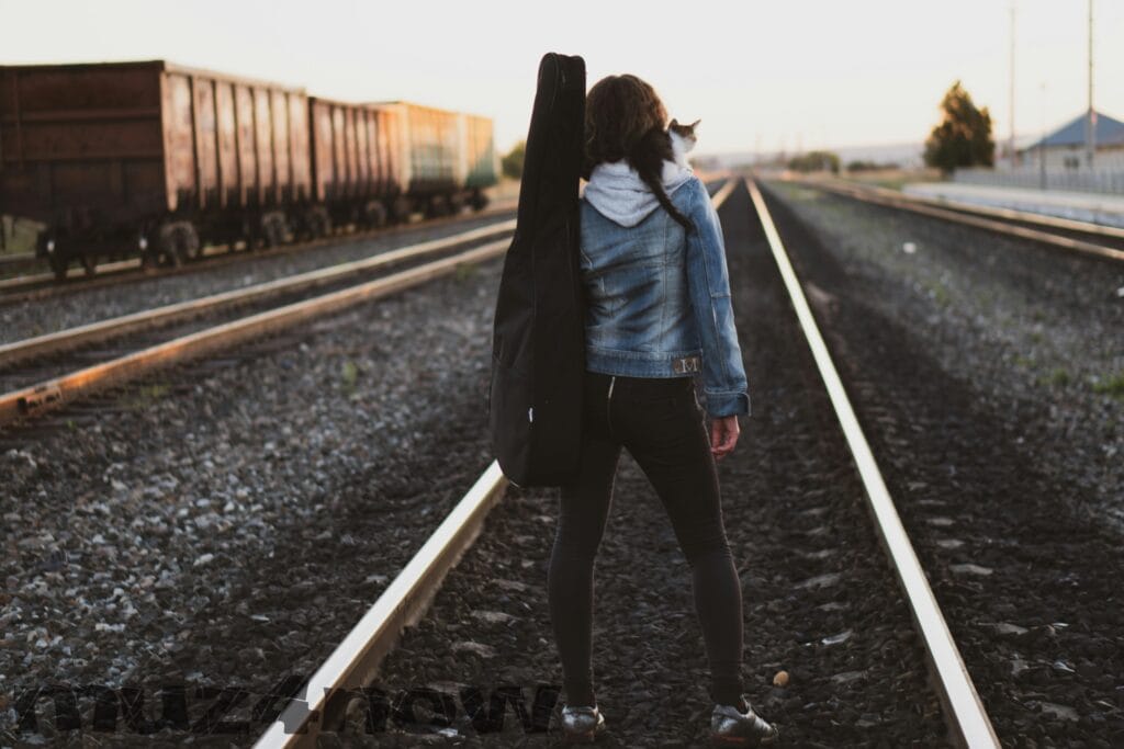 A person with a guitar bag strapped on their shoulder facing in the other direction, looking down a train track that seems to go on forever. There is a kitten on the person's shoulder. Visit https://www.pexels.com/@ruslan/ for this photographer's work.