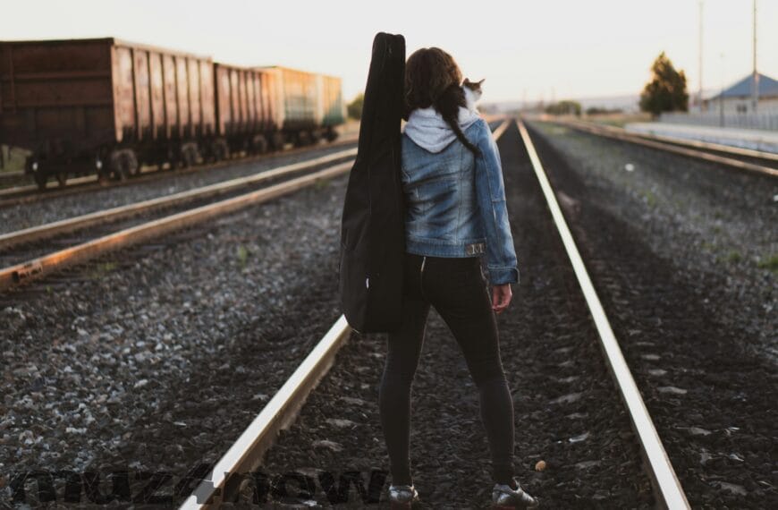 A person with a guitar bag strapped on their shoulder facing in the other direction, looking down a train track that seems to go on forever. There is a kitten on the person's shoulder. Visit https://www.pexels.com/@ruslan/ for this photographer's work.