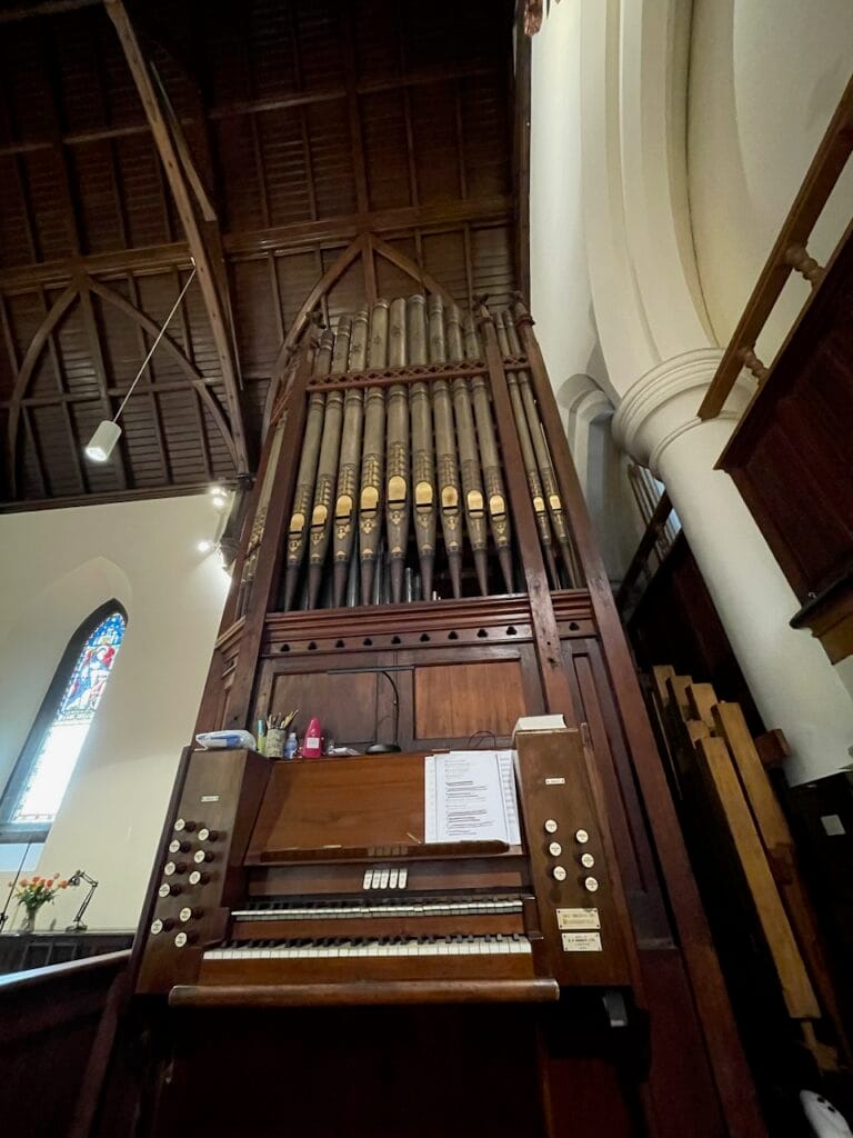 A pipe organ with 2 manuals (keyboards) and 16 visible stops. The pipes are tall above the keyboards. There is a stained glass window with bright light coming through it to the left of the organ console. On the right side of music stand built into the console is a printed form.