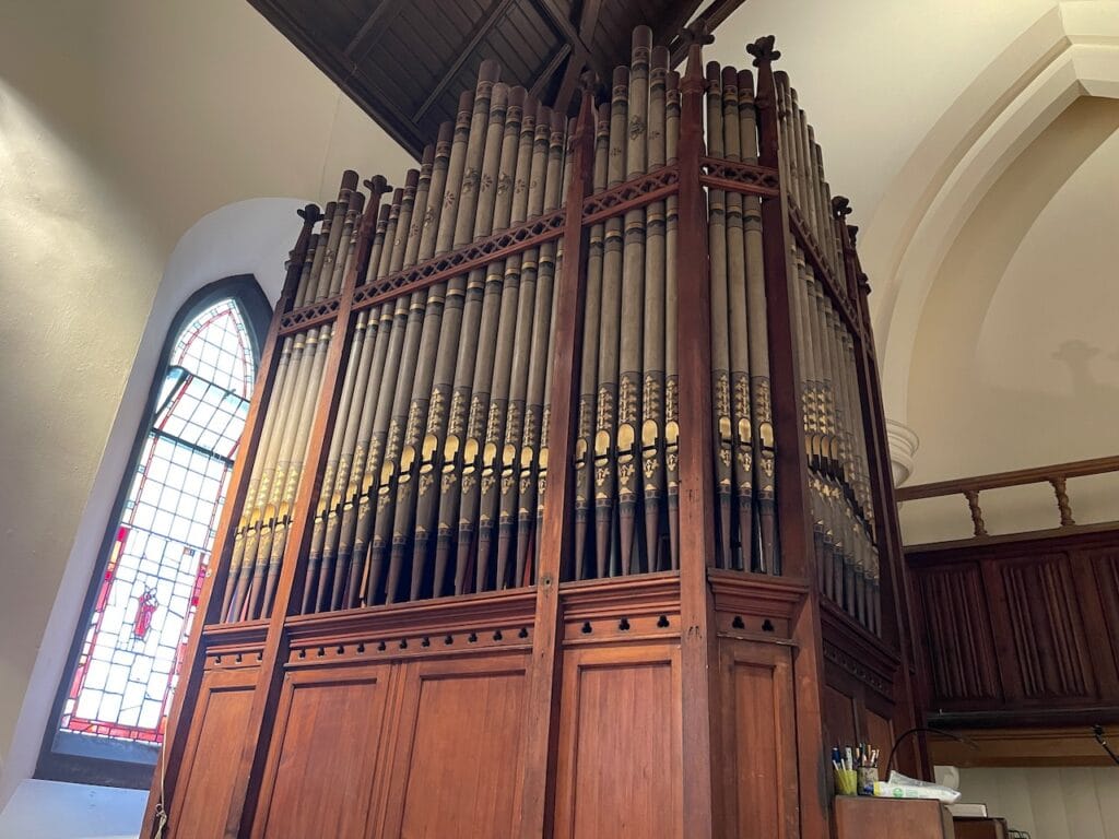 An array of pipes as part of a pipe organ. The pipes are tall and divided into 5 uneven sections.
There is a brightly illuminated stained glass window on the left end of the pipe array.