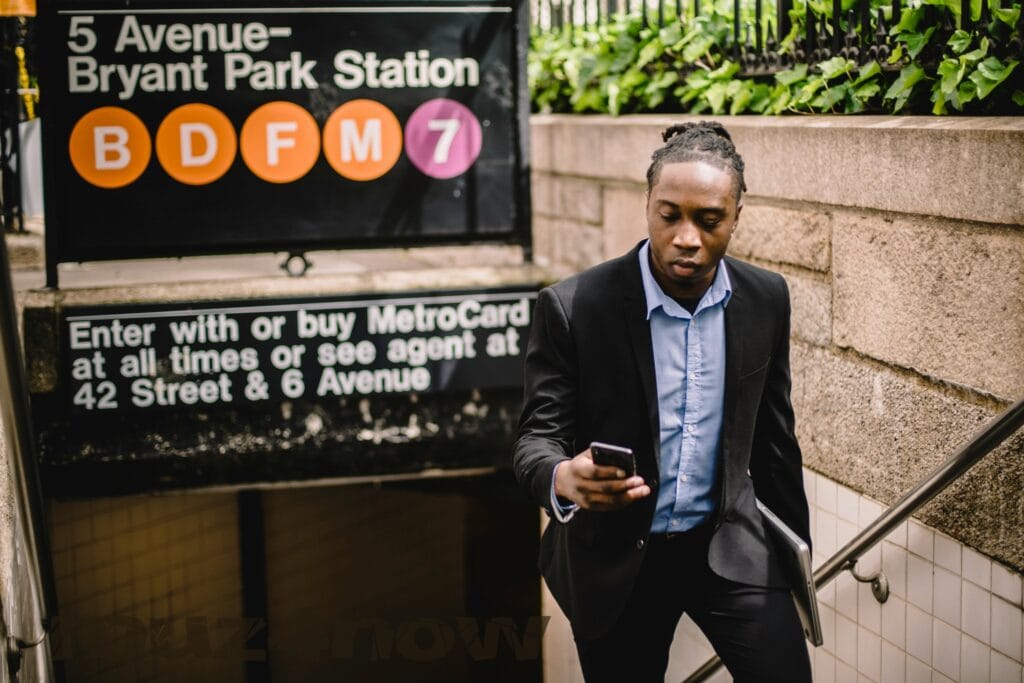 Numbers and letters on a subway sign fill most of this photo of someone wearing a dark jacket and looking at their phone as they come up a stairway.