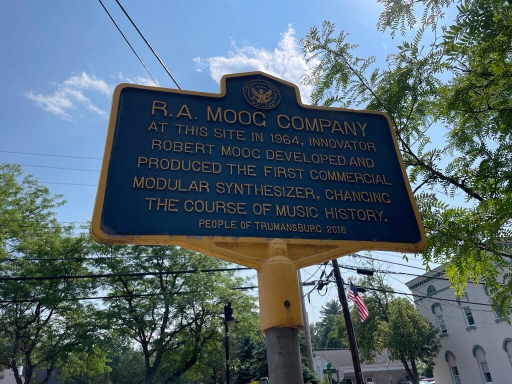 A blue sign with yellow writing:
"R.A. MOOG COMPANY
At this site in 1964, innovator Robert Moog developed and produced the first commercial modular synthesizer, changing the course of music history.
PEOPLE OF TRUMANSBURG 2016"