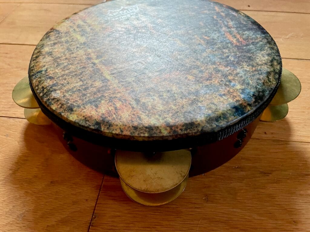 A riqq hand drum with brass jingles on a wooden floor. The head on this rig is multi-colored brown, black, muted blue, and red.
