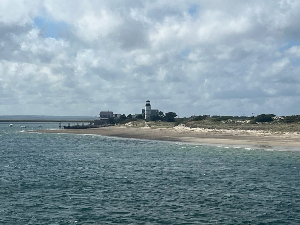 Under a cloudy sky, a lighthouse is prominent among a few other buildings just beyond a smooth, sandy beach.
