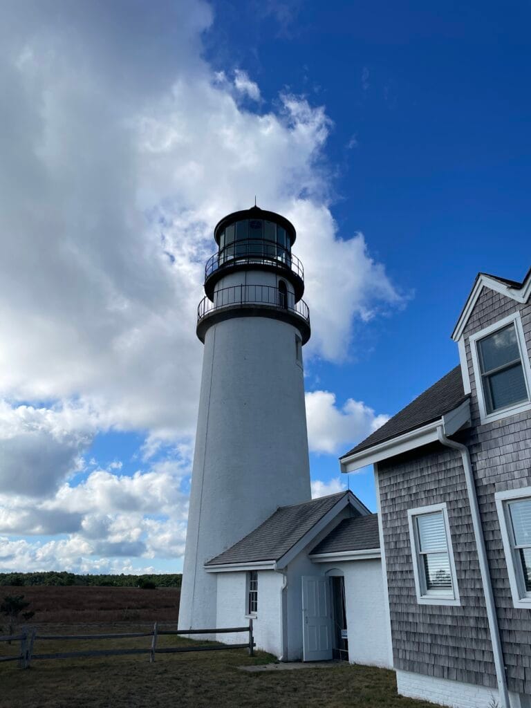 A white lighthouse with a mixed sky of blue and clouds. To the right of the lighthouse is a small building with an A-shaped roof and then a larger building with rough wooden shingles on its walls. The buildings each have windows. There is a wooden fence with 2 vertical beams extending from the smaller building.