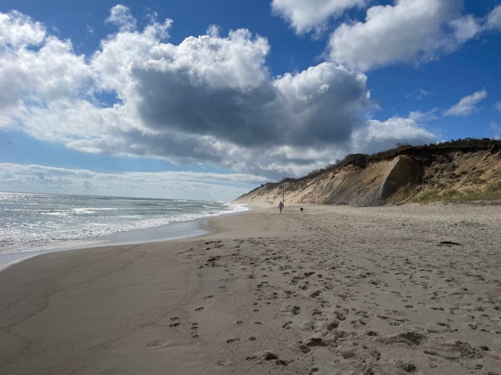 An ocean beach with sand dunes behind it and a partly cloudy sky above it.