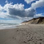 An ocean beach with sand dunes behind it and a partly cloudy sky above it.