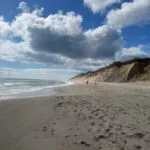 An ocean beach with sand dunes behind it and a partly cloudy sky above it.