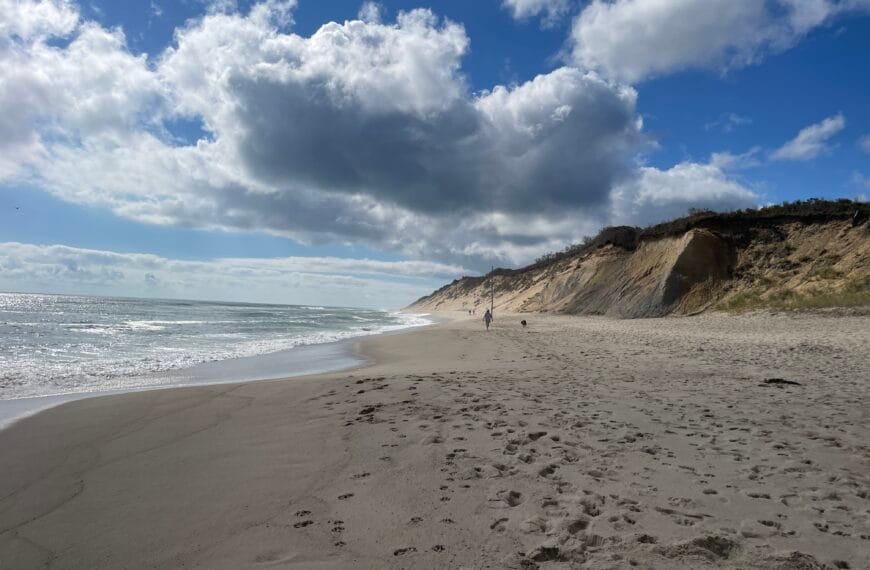 An ocean beach with sand dunes behind it and a partly cloudy sky above it.