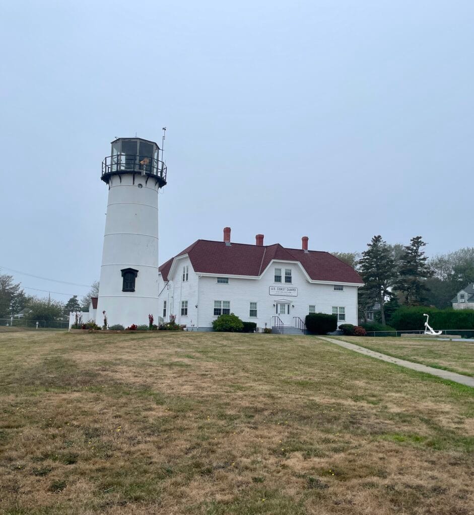 A white lighthouse and building beyond a lawn that is mostly brown grass. The building has a red shingle roof and 3 chimneys. There is a white anchor on the lawn and to the right of the building. The sky is solid grey.