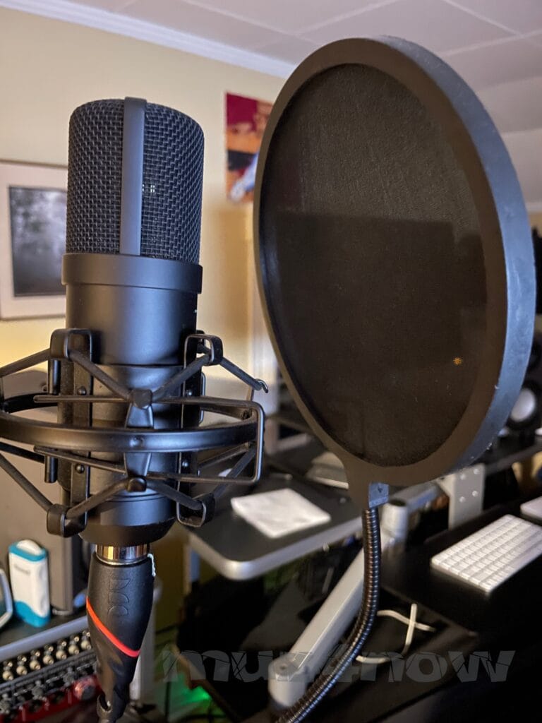 A studio recording microphone with a pop screen in front of it fills the foreground. A computer, speakers, wall art, and other objects are blurred in the background.