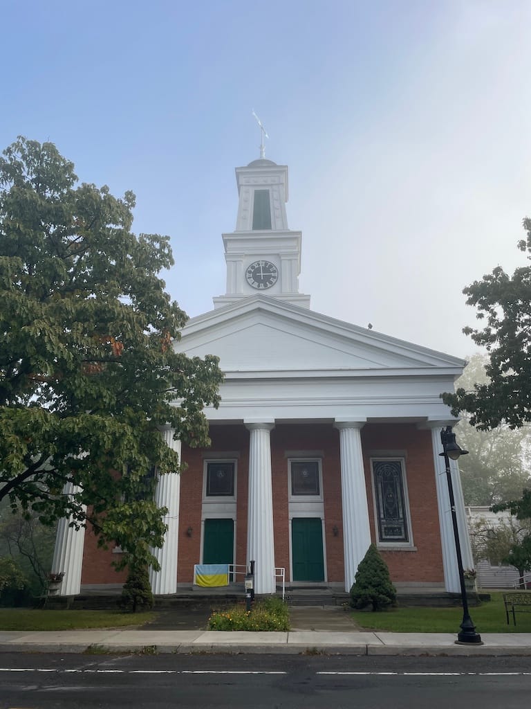 The front of a church with pillars along a front porch and a steeple with a clock on it. There is a Ukrainian flag symbolizing people fighting for freedom on the handicap ramp.