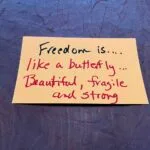 Handwritten in black and red marker on a peach-colored index card: Freedom is.... like a butterfly ... Beautiful, fragile and strong