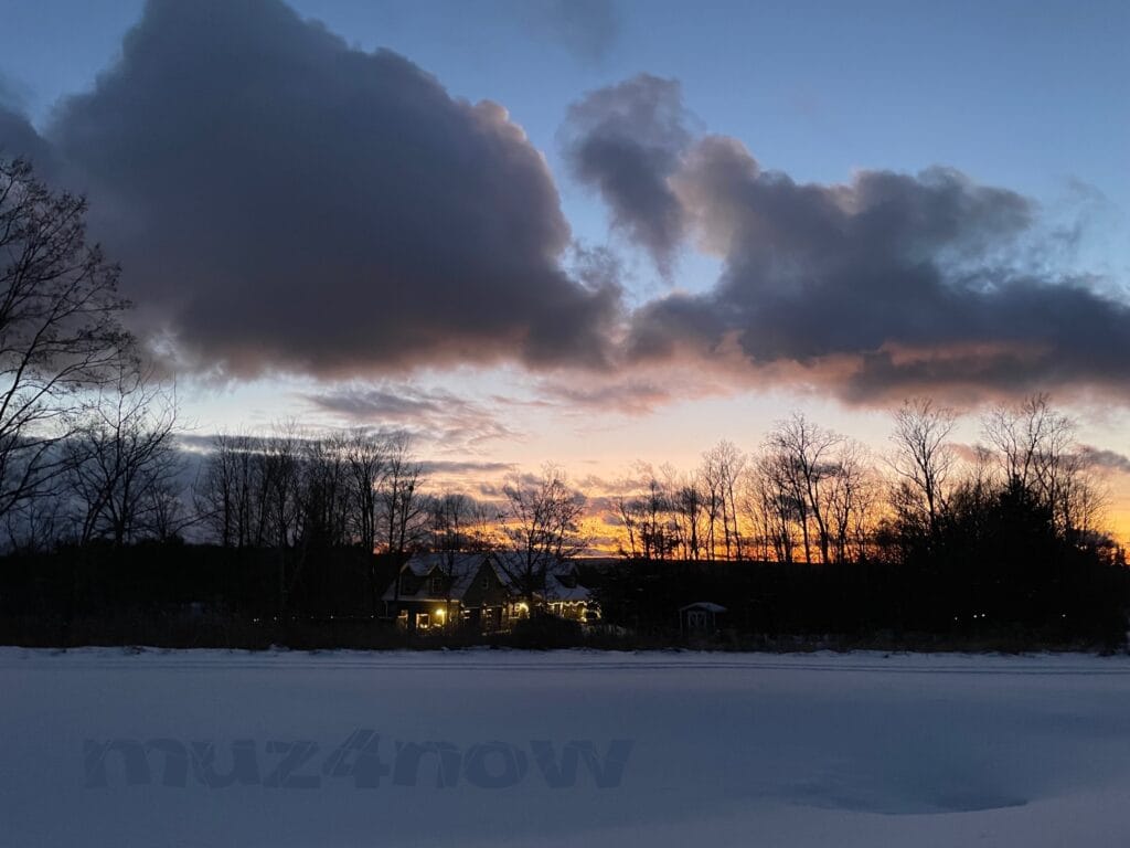 The sunset through deep gray winter clouds with a house lit up with holiday lights nestled in a grove of trees. There is snow covering the ground in the foreground.