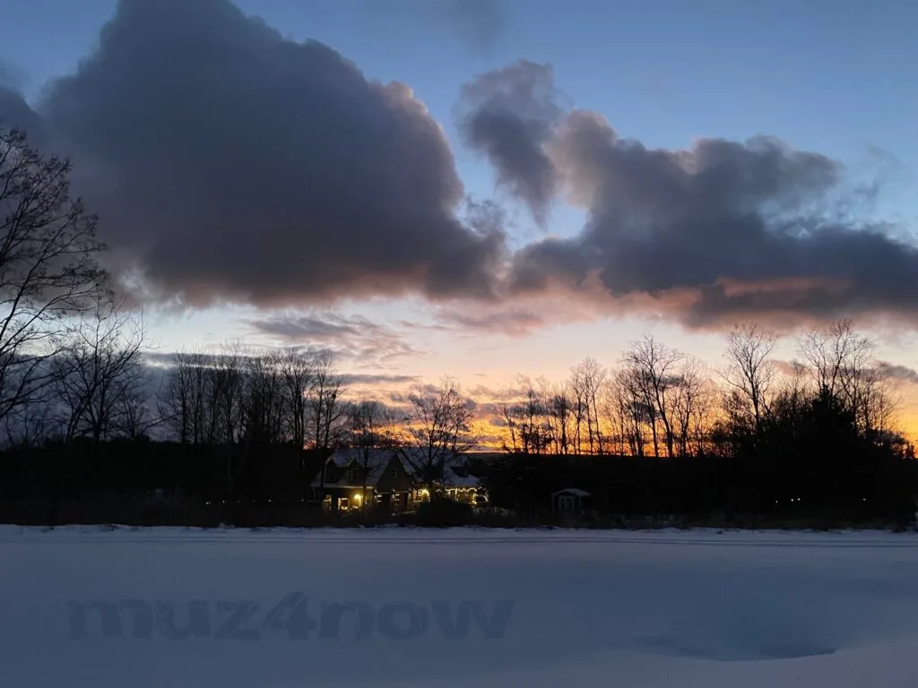 The sunset through deep gray winter clouds with a house lit up with holiday lights nestled in a grove of trees. There is snow covering the ground in the foreground.