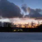 The sunset through deep gray winter clouds with a house lit up with holiday lights nestled in a grove of trees. There is snow covering the ground in the foreground.