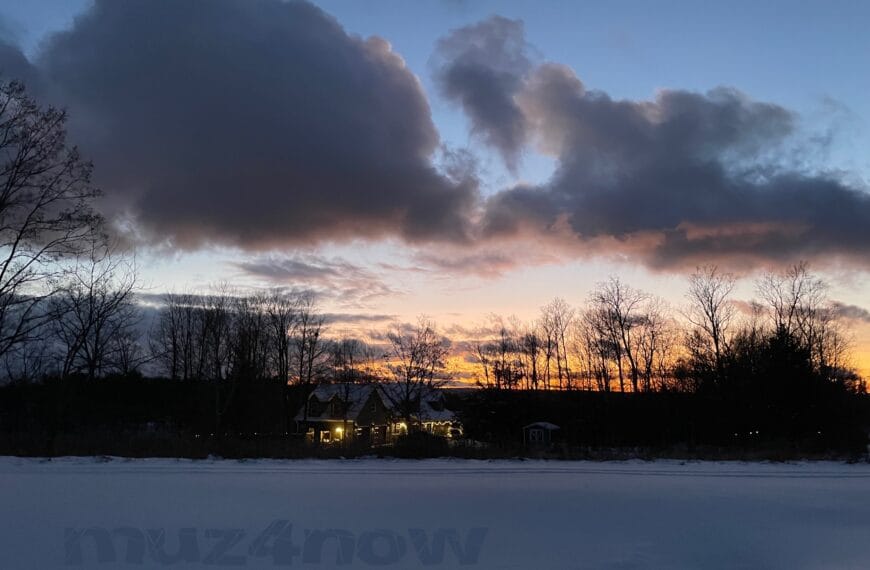 The sunset through deep gray winter clouds with a house lit up with holiday lights nestled in a grove of trees. There is snow covering the ground in the foreground.