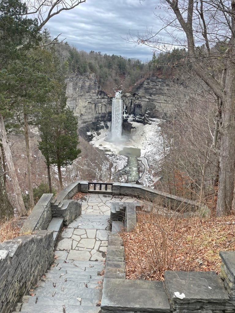 Out beyond a stone overlook is a tall waterfall, crashing down a gray rocky cliff. As if framing the waterfall, there are leafless and evergreen trees on both sides of the foreground.
Mid-winter wonders in the Finger Lakes.