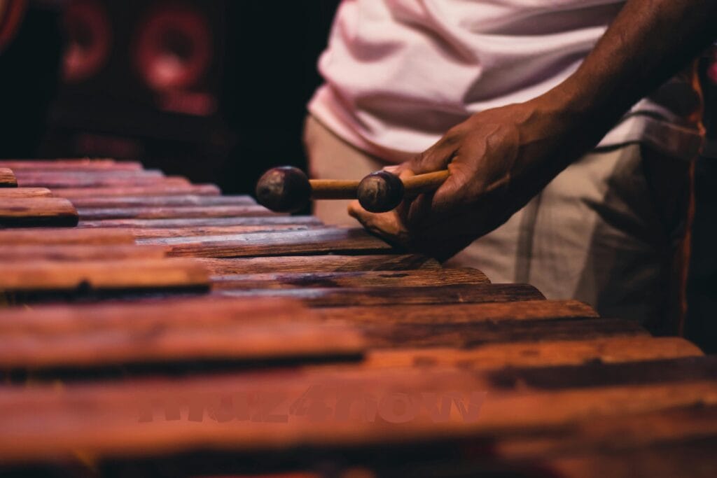 Rough-carved marimba keys fill the frame with someone holing two mallets in the background.
