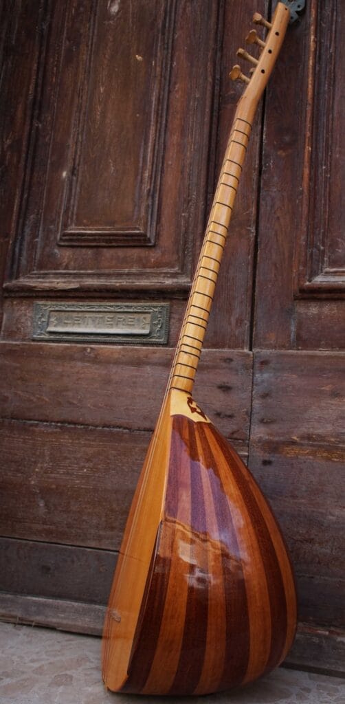 Leaning against a door is a stringed instrument called the saz. It has a large resonating chamber (called a "bowl") resting on the floor. This chamber has alternating darker and lighter reddish tones. Both the instrument and the door are made of wood.
For a blog post highlighting musical instruments seen in the West as "foreign".