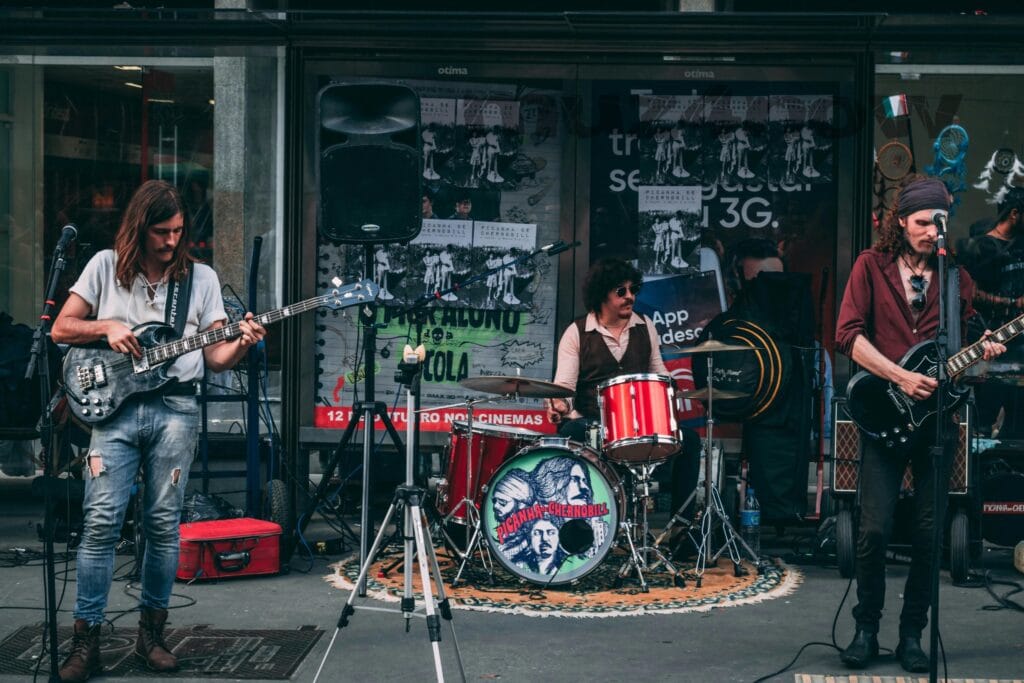 A photo of a 3-piece rock band with bass player looking down at his guitar on the left, drummer on a multi-piece drum kit in the center, and electric guitar player on the right. They are on a stage with equipment around them. The photo is by https://www.pexels.com/@athena/