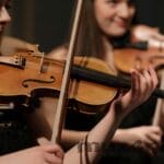 Two violins being bowed by players with bare forearms. The violinist in the background is smiling. The photo is by https://www.pexels.com/@cottonbro/