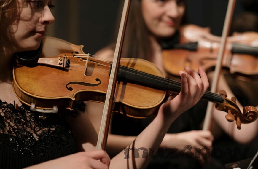 Two violins being bowed by players with bare forearms. The violinist in the background is smiling. The photo is by https://www.pexels.com/@cottonbro/