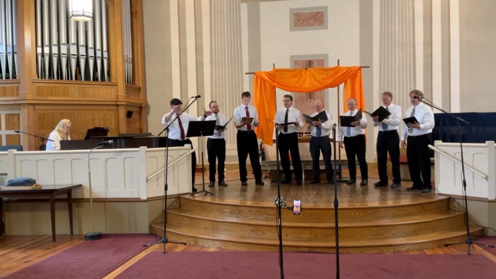 A group of 8 singers --the one on the far left is conducting -- and a pianist performing. Hopefully they don't experience the blahs after the gig.