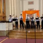 A group of 8 singers --the one on the far left is conducting -- and a pianist performing. Hopefully they don't experience the blahs after the gig.