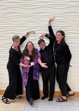 Four people dressed mostly in black stand in front of a white wave wall making unusual shapes with their arms and hands.