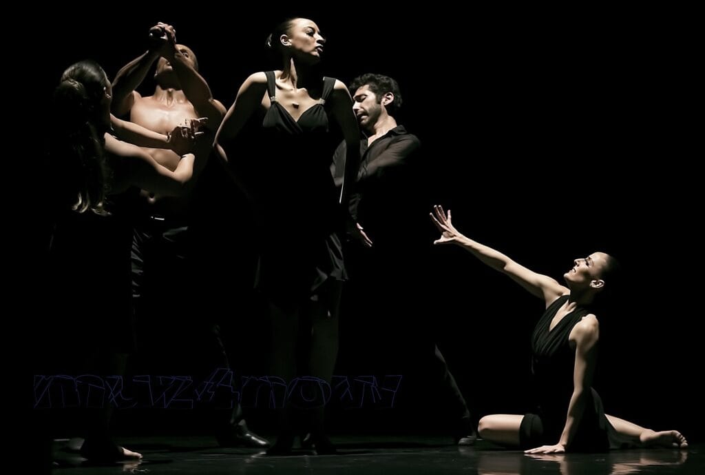 Five dancers in a live performance on a dark stage. One dancer is on the floor reaching out to the other four.