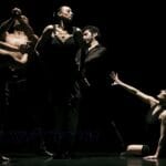 Five dancers in a live performance on a dark stage. One dancer is on the floor reaching out to the other four.