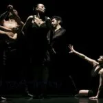 Five dancers in a live performance on a dark stage. One dancer is on the floor reaching out to the other four.