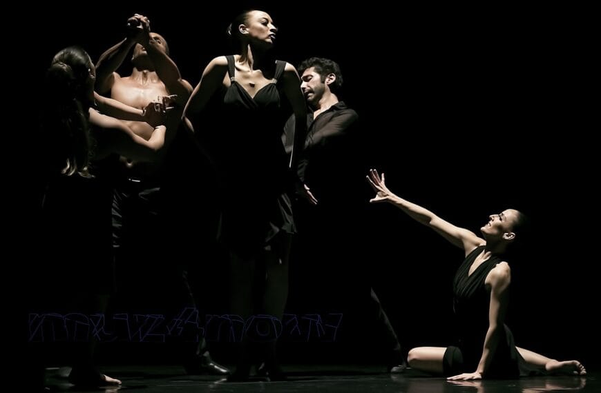 Five dancers in a live performance on a dark stage. One dancer is on the floor reaching out to the other four.