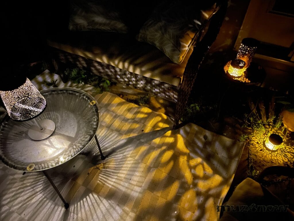 An outdoor love bench and round table are surrounded by a variety of solar-powered lamps. The lamps are shining yellow lights in the shapes cut in their sides.