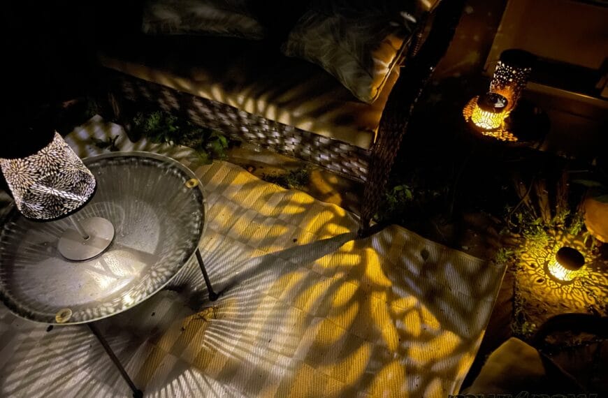 An outdoor love bench and round table are surrounded by a variety of solar-powered lamps. The lamps are shining yellow lights in the shapes cut in their sides.