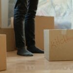 Legs of someone in jeans and wearing socks stand in the middle of some cardboard boxes. One box has "kitchen" written on it. Another one has "recording studio" printed in red permanent marker.