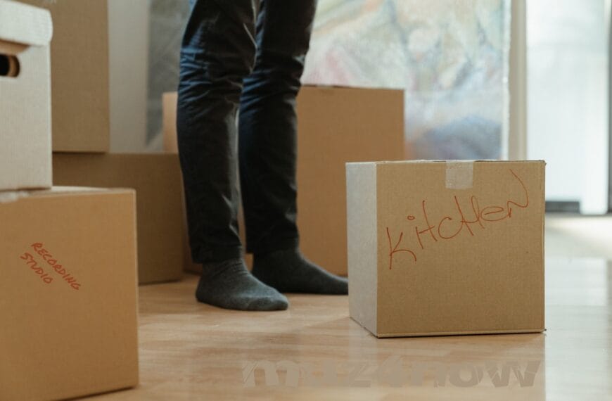 Legs of someone in jeans and wearing socks stand in the middle of some cardboard boxes. One box has "kitchen" written on it. Another one has "recording studio" printed in red permanent marker.