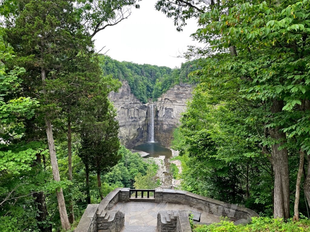 A tall waterfall pours down a grey rock face. All around the rocks are deep, green forests.