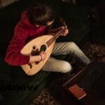 Someone seated on a couch, playing taqsim on an oud. They are wearing a red sweatshirt and white pants. Their hair is black and tied back. The perspective of the image is from above. The photo is by https://www.pexels.com/@abedalbaset/