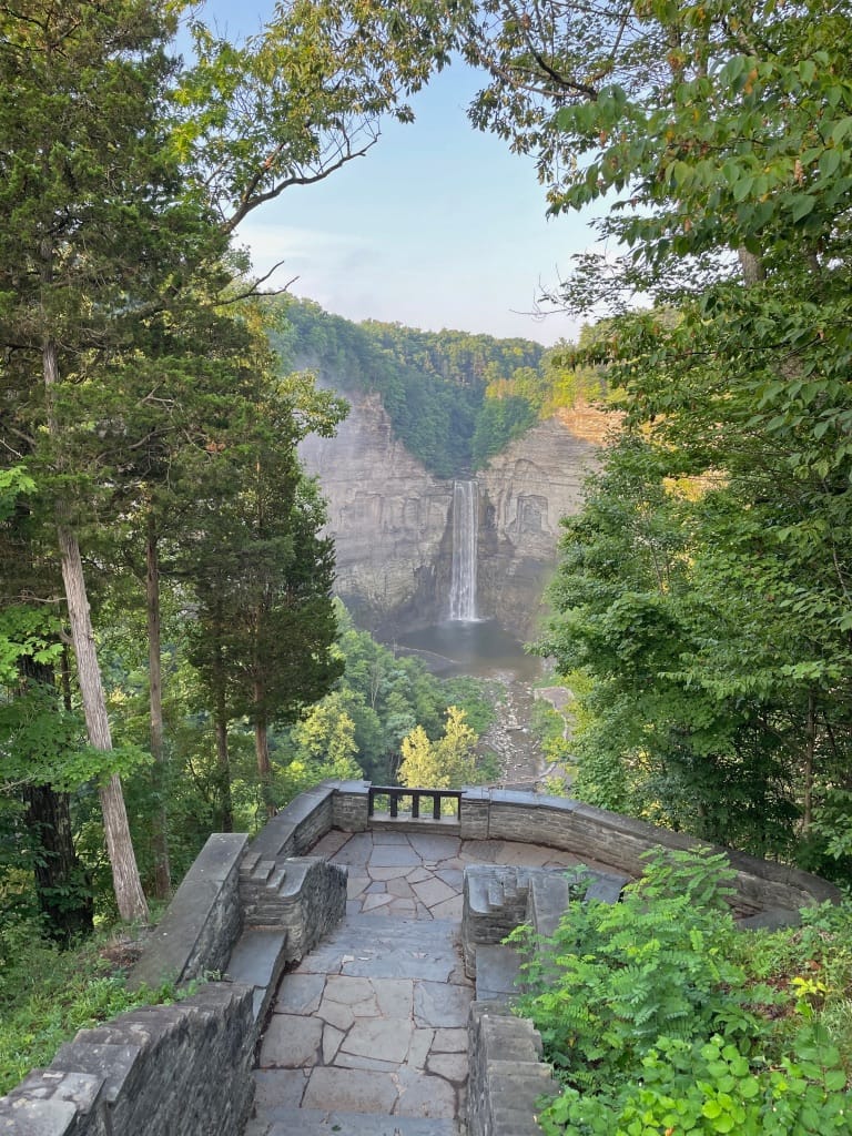 A tall waterfall is tumbling down a rocky cliff. The falls are framed by a rock overlook and bright green forest in the foreground.